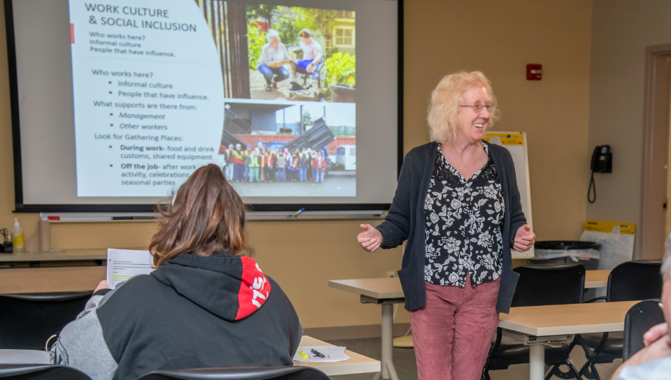 Coordinator of Access to Integrated Employment Vicki Ferrara leads a session of Supporting Meaningful Employment in the training room at the Sherlock Center. On the screen behind her is a PowerPoint that reads Work Culture and Social Inclusion.