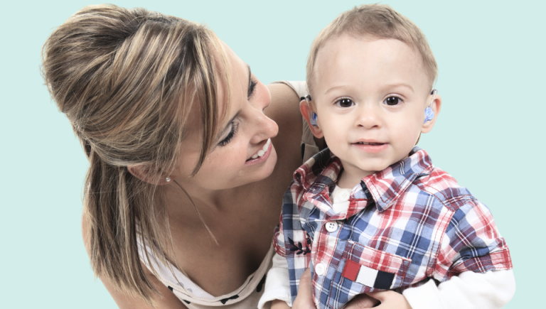 A woman holds a toddler boy in a blue and red plaid shirt wearing hearing aids.