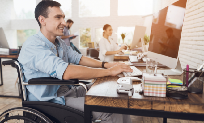 A man who uses a wheelchair is pictured at a computer in an office with his co-workers in the background.