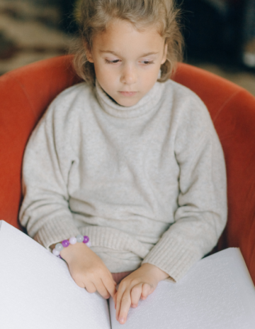 A young girl sits in a chair reading a braille book.