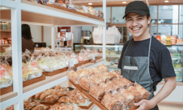 A young man wearing a hat and apron holds a tray of baked goods to restock the shelves at a bakery.