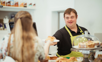 A man with an intellectual disability serves a scone to a woman in the café where he works.