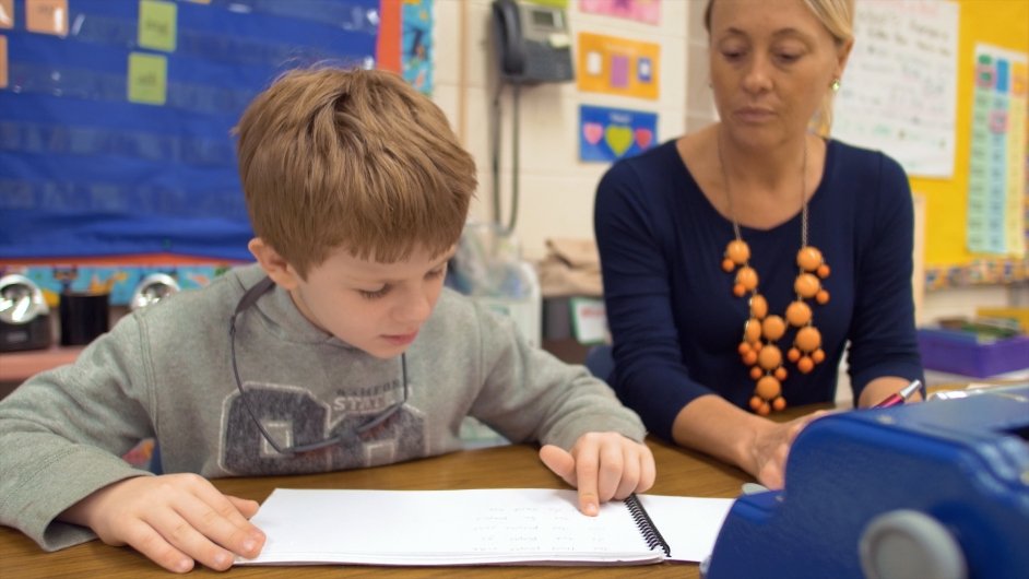 A visually impaired student reads a braille book as a teacher sits next to him.
