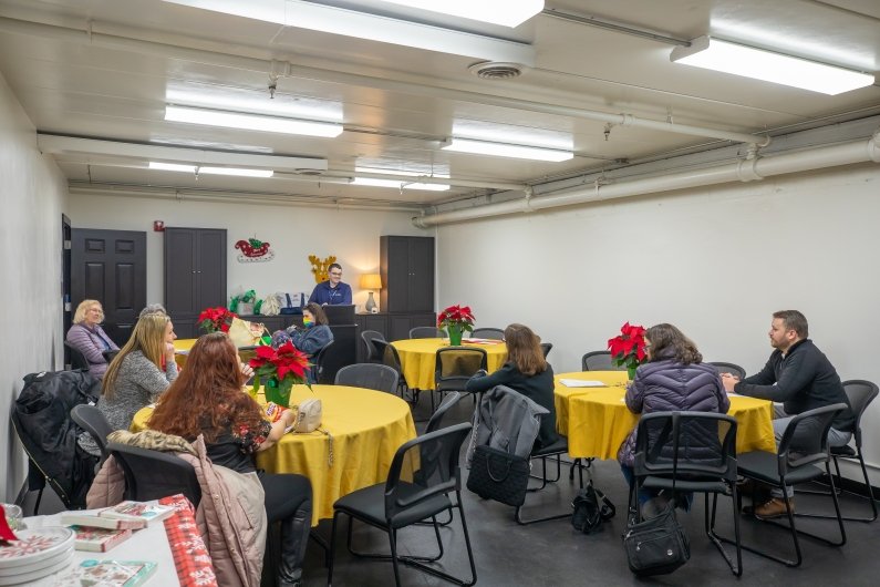 This photo shows the group from the back of the room with Sentinels facilitator Conor O’Brien leading the meeting from a lectern in the front. The room is decorated with poinsettias and members are sitting at round tables. 
