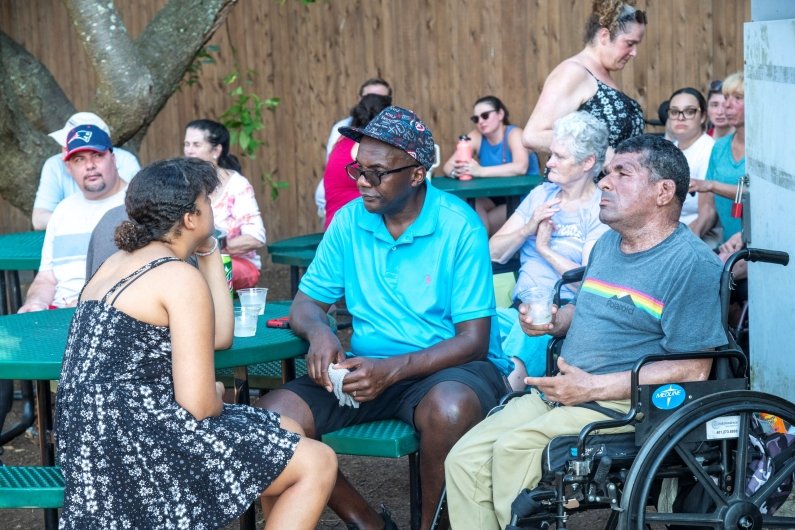 Three guests are seated at a picnic table with others in the background.