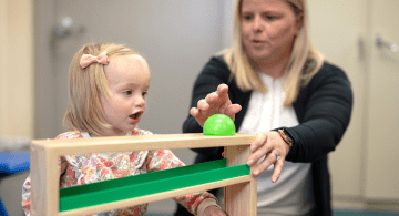 An early intervention specialists works with a blonde toddler girl playing with a small green ball on a toy with ramps.