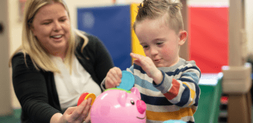 An early intervention specialist works with a young boy putting plastic coins in a piggy bank.
