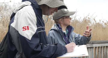 Two people are outdoors; one is gesturing and the other is making a list.