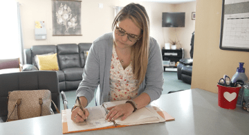 A woman writes in a log book on a desk.