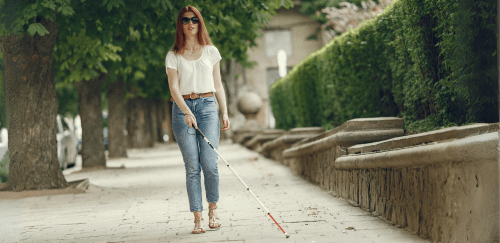 A young blind woman uses a white cane while walking on a sidewalk.