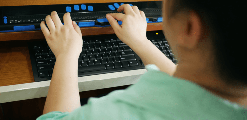 Rear view of a person working with a computer keyboard and braille display as a technology assistive device for people with visual impairment.