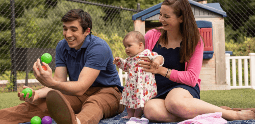 A family is sitting on a blanket at the playground. The father is juggling colorful, small balls while the mother helps a baby stand to watch.