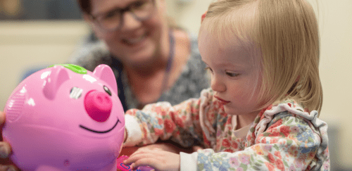 A toddler girl plays with a pink piggy bank as an early intervention professional looks on.