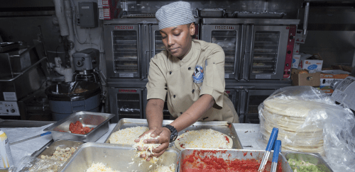 A young woman makes pizza in a commercial kitchen.