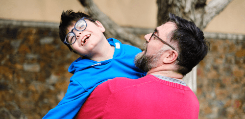 A father wearing a red shirt holds and plays with his son, who is wearing  glasses and a blue shirt.