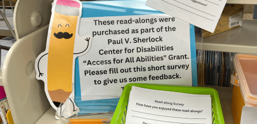 A sign at the Jesse M. Smith Memorial Library says "These read-alongs were purchased as part of the Paul V. Sherlock Center on Disabilities "Access for All Abilities" Grant. Please fill out this short survey to give us some feedback." Next to it are a pile of survey forms and pencils.