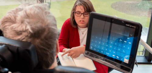 An adult in a red sweater shows a notebook to an adult in a blue shirt using a wheelchair with augmentative and alternative communication (AAC) device, a tablet that helps someone with a speech or language impairment to communicate.