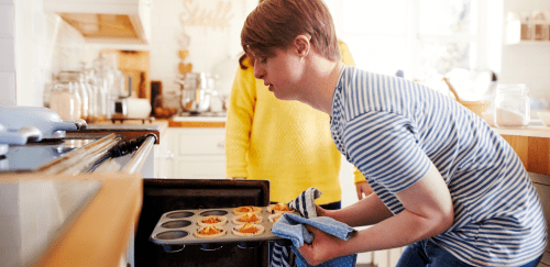 A young man with Down syndrome takes a muffin tin out of the oven. 