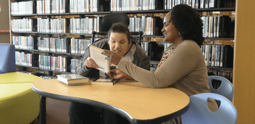 A direct support professional reads with a woman in the library.