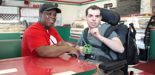 A direct support professional sits with a man in a wheelchair in a diner.