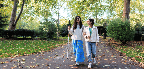 Two women walk among fallen leaves. One is using a white cane.