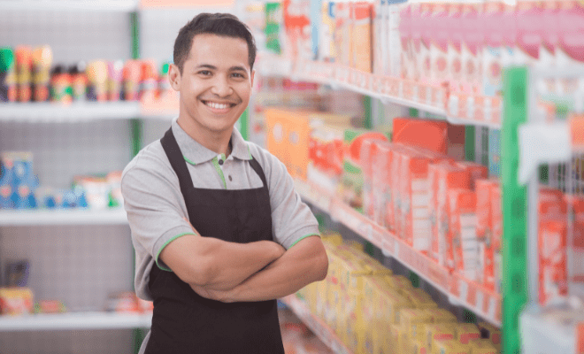 A smiling young man stands in the aisle of a grocery store where he works.