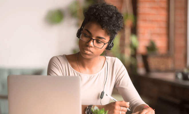 A woman wearing headphones takes notes as she participates in a Zoom meeting on a laptop.